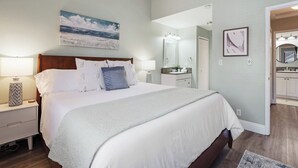 Une chambre à coucher avec un grand lit, des lampes de chevet, un tableau représentant une plage et une salle de bain visible à travers une porte ouverte.