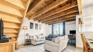 Een ruime woonkamer met een hoge plafond en zichtbare houten balken, een zithoek, een eettafel en een televisie.
