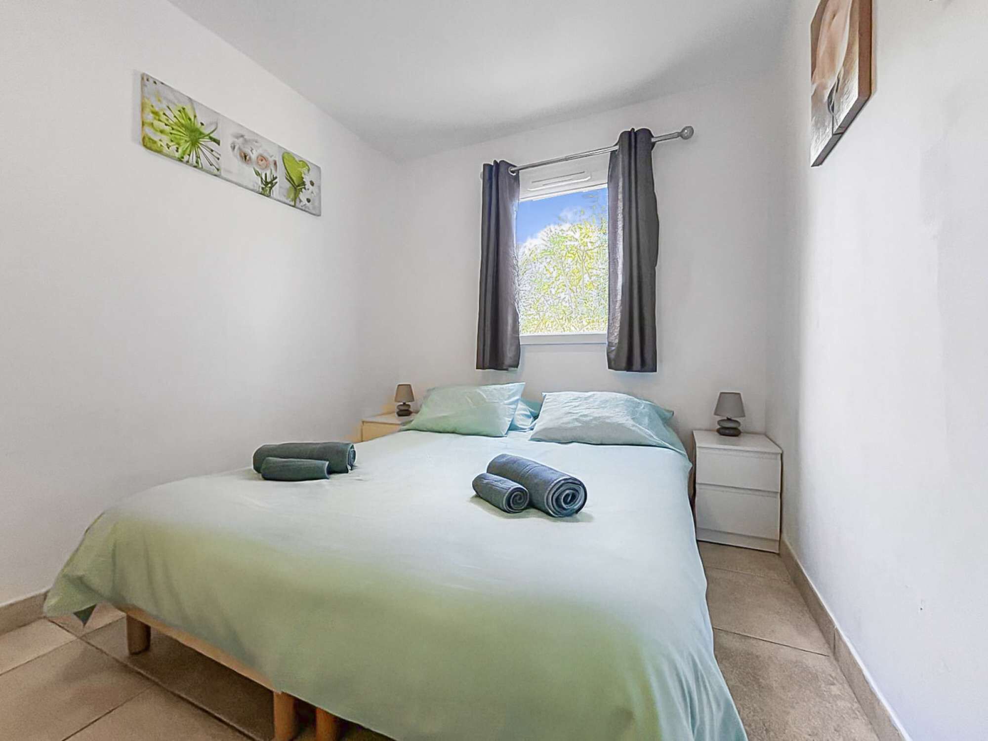 A bedroom with a bed, two bedside tables, a window with curtains, and a wall-mounted picture.