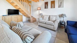 Een woonkamer met een bank, een blauwe fauteuil, een televisie op een houten kast, een trap en drie wanddecoraties met bladmotieven.