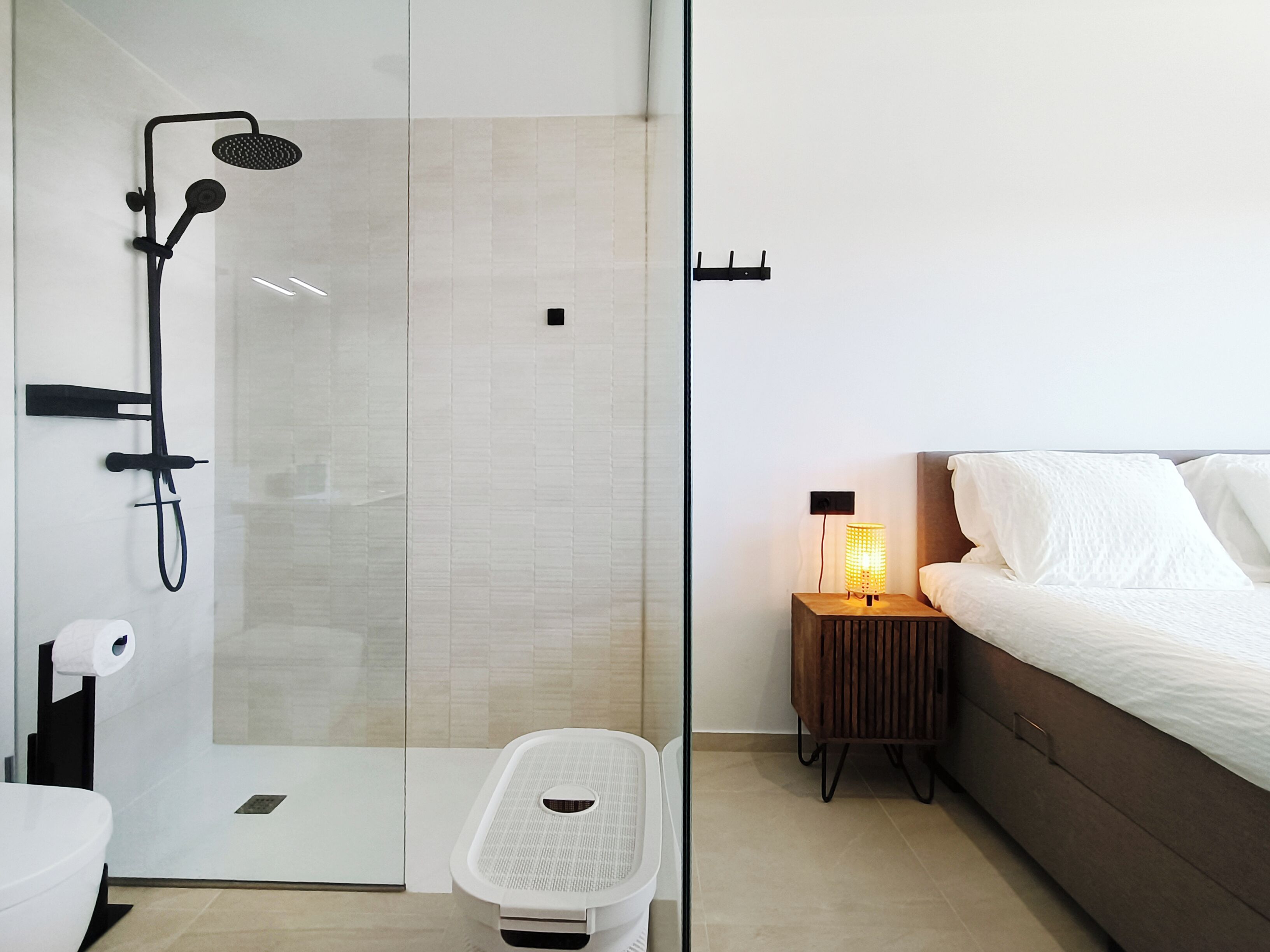 Una habitación de hotel moderna con cabina de ducha de vidrio, una mesita de noche con lámpara y una cama con ropa blanca.