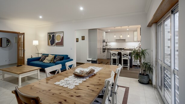 A modern living room with a wooden dining table, white chairs, a blue sofa, and a kitchen in the background. - Carcavelos Bright Modern Apartment by Homing (Cascais)