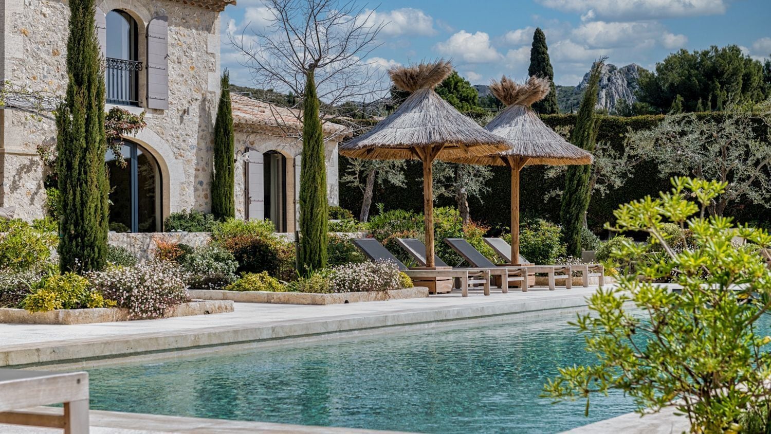 Un espace piscine avec des chaises longues, un bâtiment en pierre et des parasols en chaume.