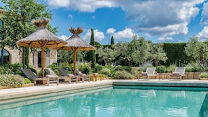 A pool area with lounge chairs and thatched umbrellas, surrounded by greenery and a stone building. - Maison Sanctuaire - Five Bedroom Villa, Sleeps 10 (Maussane-les-Alpilles)