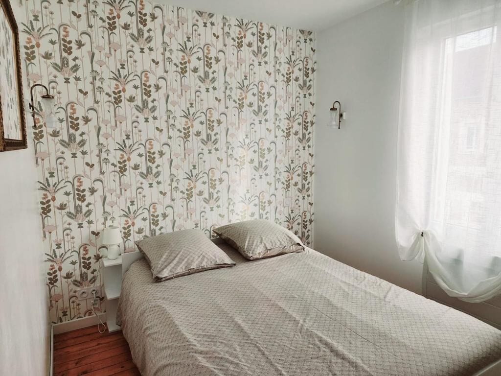 A bedroom with a floral wallpaper, a bed with two pillows, a bedside table with a lamp, and a window with sheer curtains.