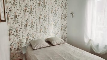 A bedroom with a floral wallpaper, a bed with two pillows, a bedside table with a lamp, and a window with sheer curtains.