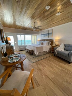 Una camera da letto con soffitto in legno, un letto, un divano grigio, un tavolo da pranzo con sedie e vista sull'oceano dalla finestra.