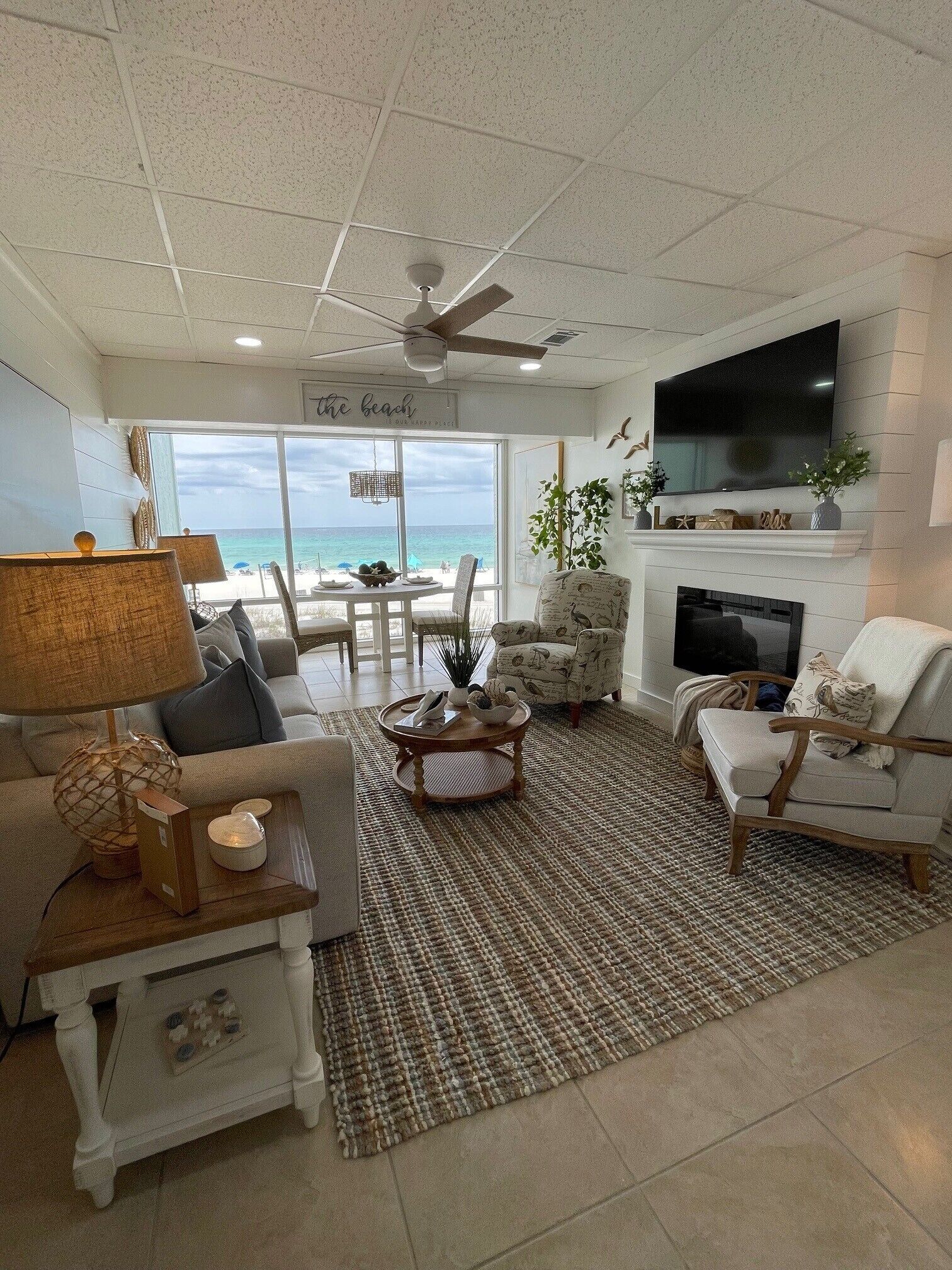 A living room with a view of the beach, a ceiling fan, a fireplace, and a television.
