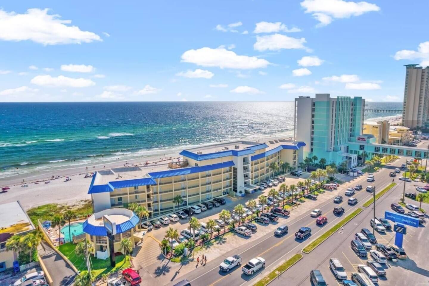 A hotel with a pool, parking lot, and a view of the beach and ocean.