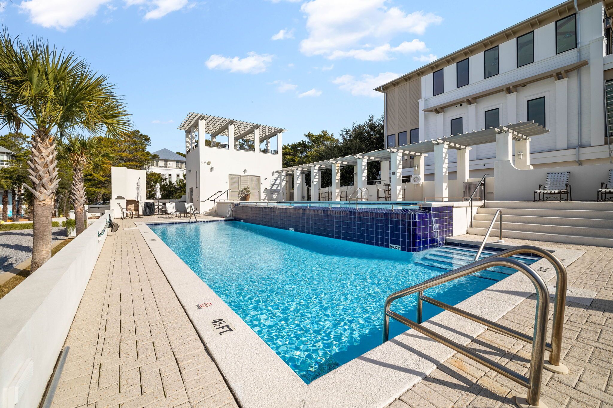 En swimmingpool med ett kaklat poolområde, en pergola och en byggnad med en vit fasad.