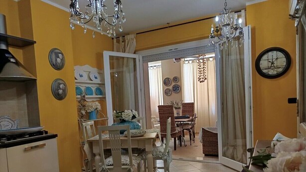 Una cucina con un piano cottura, un tavolo da pranzo, sedie e un lampadario. Si vede un orologio appeso al muro e una porta che dà su un'altra stanza.