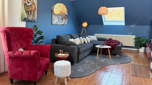 Un salon moderne avec un fauteuil rouge, un canapé foncé et une petite table ronde. On y trouve deux lampes suspendues et un tableau représentant un lion accroché au mur.