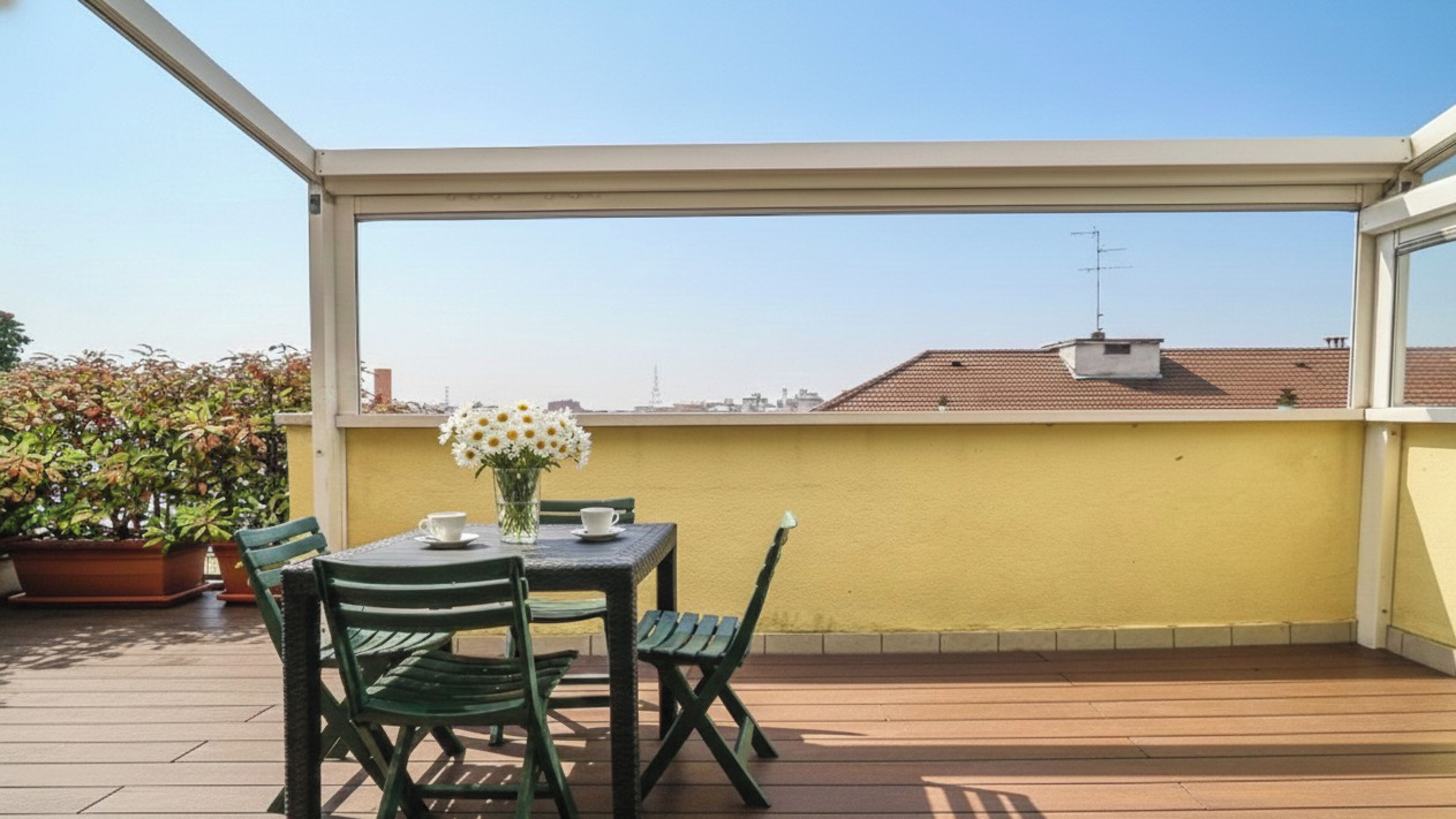 Eine überdachte Terrasse im Freien mit Tisch und Stühlen, einem Blumenstrauß und Blick auf Gebäude im Hintergrund.