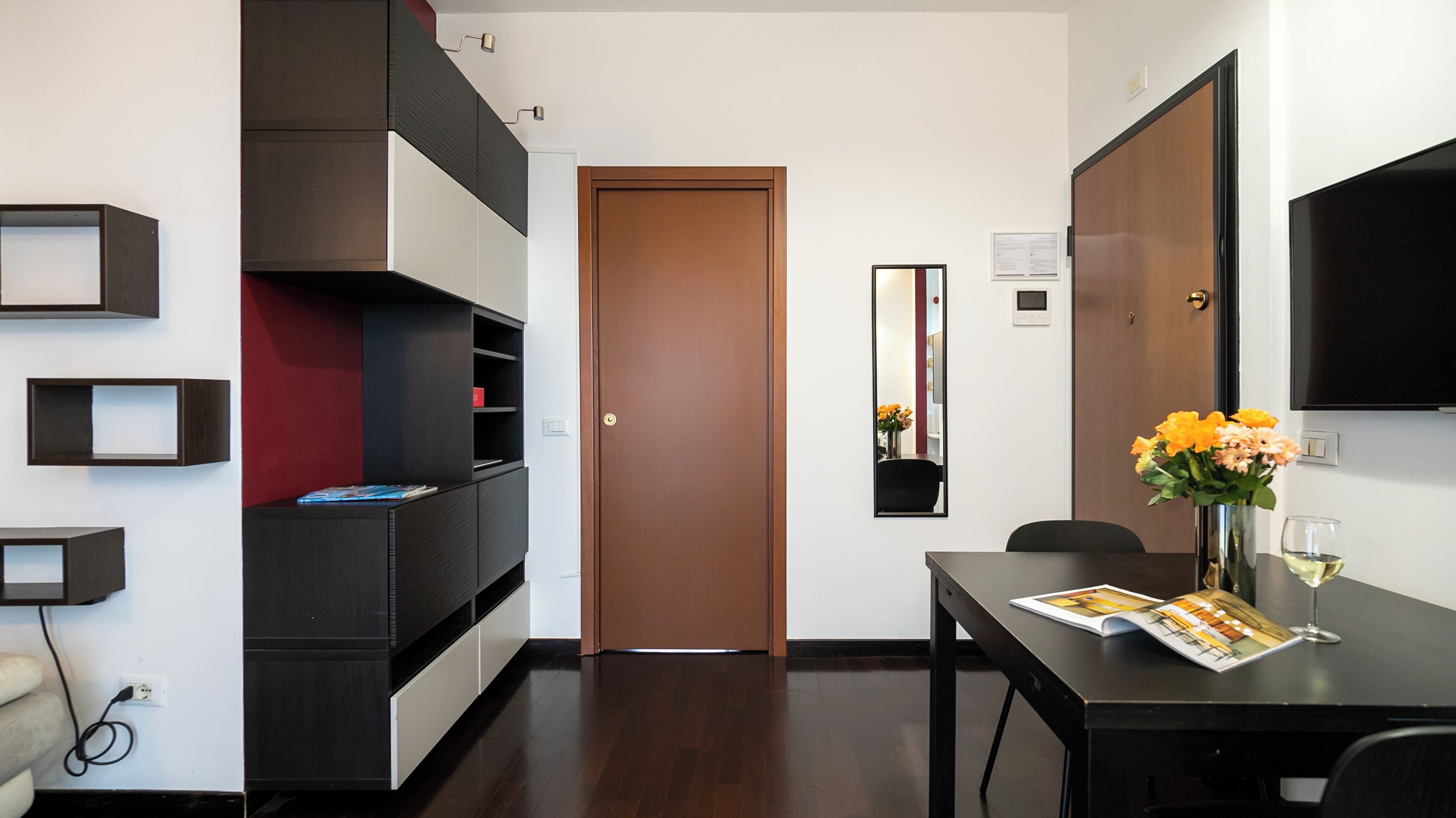 Een moderne hotelkamer met een houten deur, inbouwkasten, een flatscreen televisie en een zithoek met een tijdschrift en bloemen.