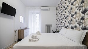 Une chambre d’hôtel avec un lit, un téléviseur à écran plat fixé au mur, une commode en bois, un radiateur et une fenêtre avec des rideaux légers.