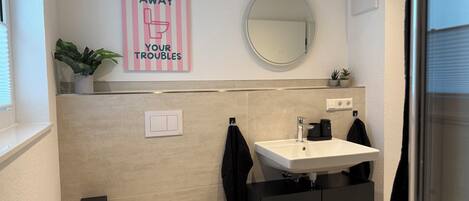 Une salle de bain avec des toilettes, un lavabo et un miroir. Au-dessus du lavabo, on peut lire l’inscription suivante : « ÉVACUEZ VOS SOUCIS DANS LES TOILETTES ».
