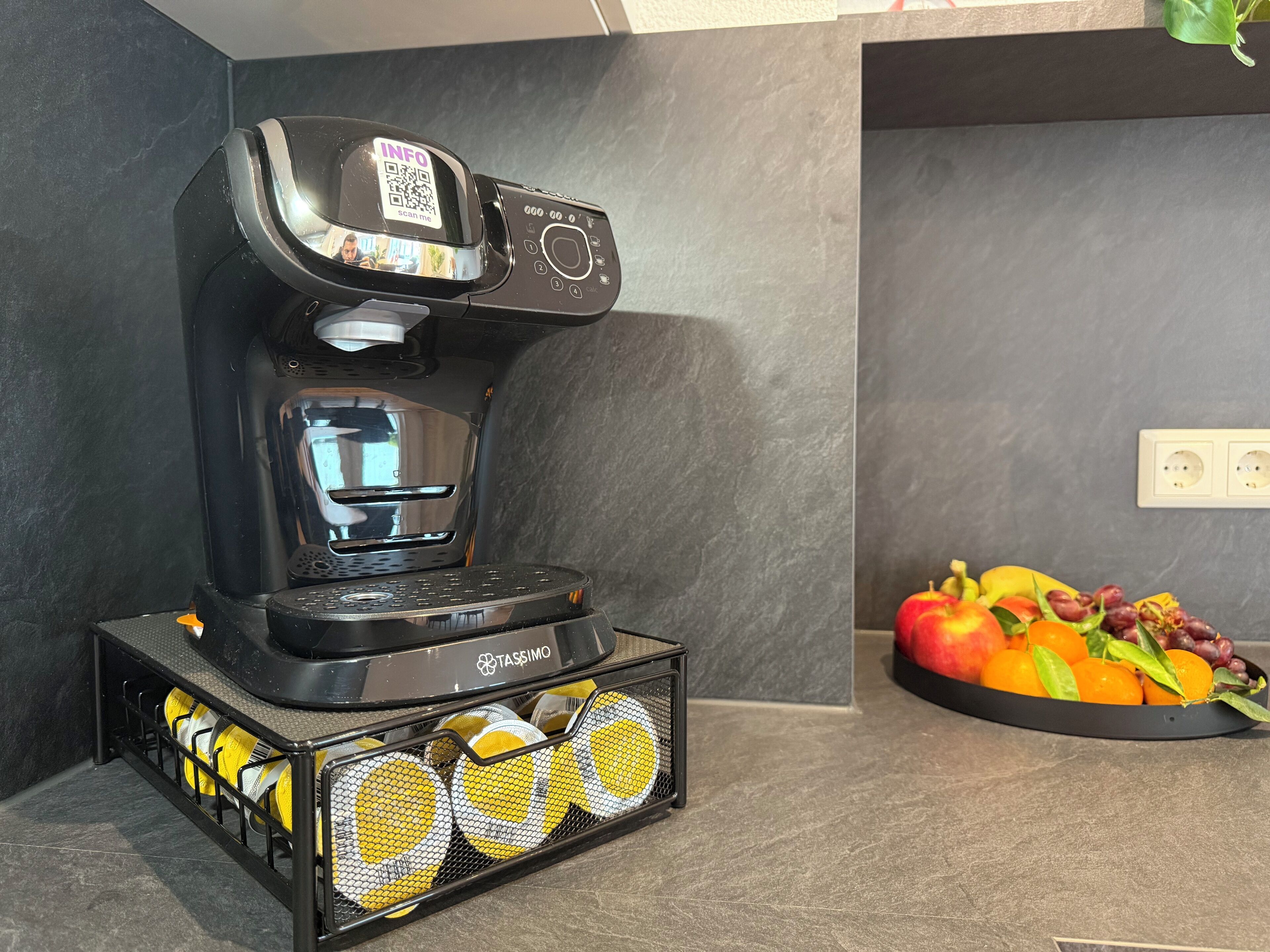 A coffee machine with a QR code, a basket of yellow and white coffee capsules, and a fruit bowl with apples, oranges, and grapes on a countertop.