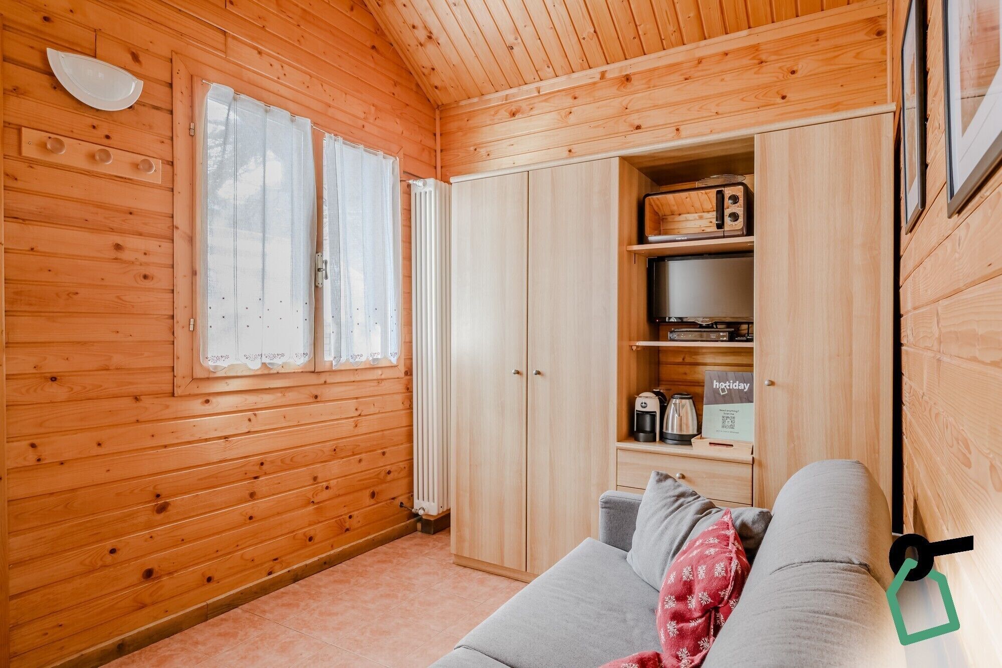 Ett mysigt rum i en stuga med träväggar, en grå soffa, ett litet bord med en vattenkokare och en hylla med en kaffebryggare.
