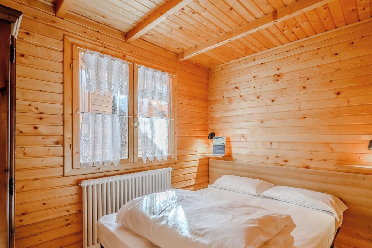 A wooden cabin room with a bed, a window with lace curtains, a radiator, and a wall-mounted light fixture.