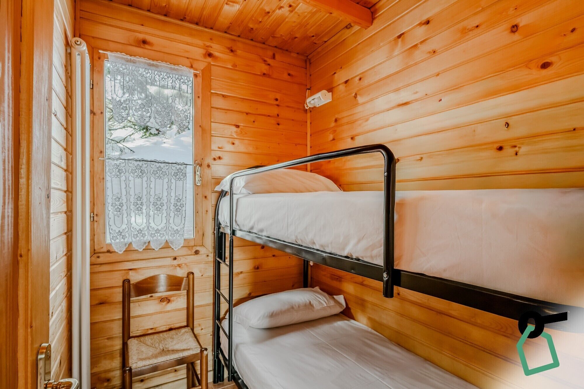 A wooden cabin room with bunk beds, a window with lace curtains, and a wooden chair.