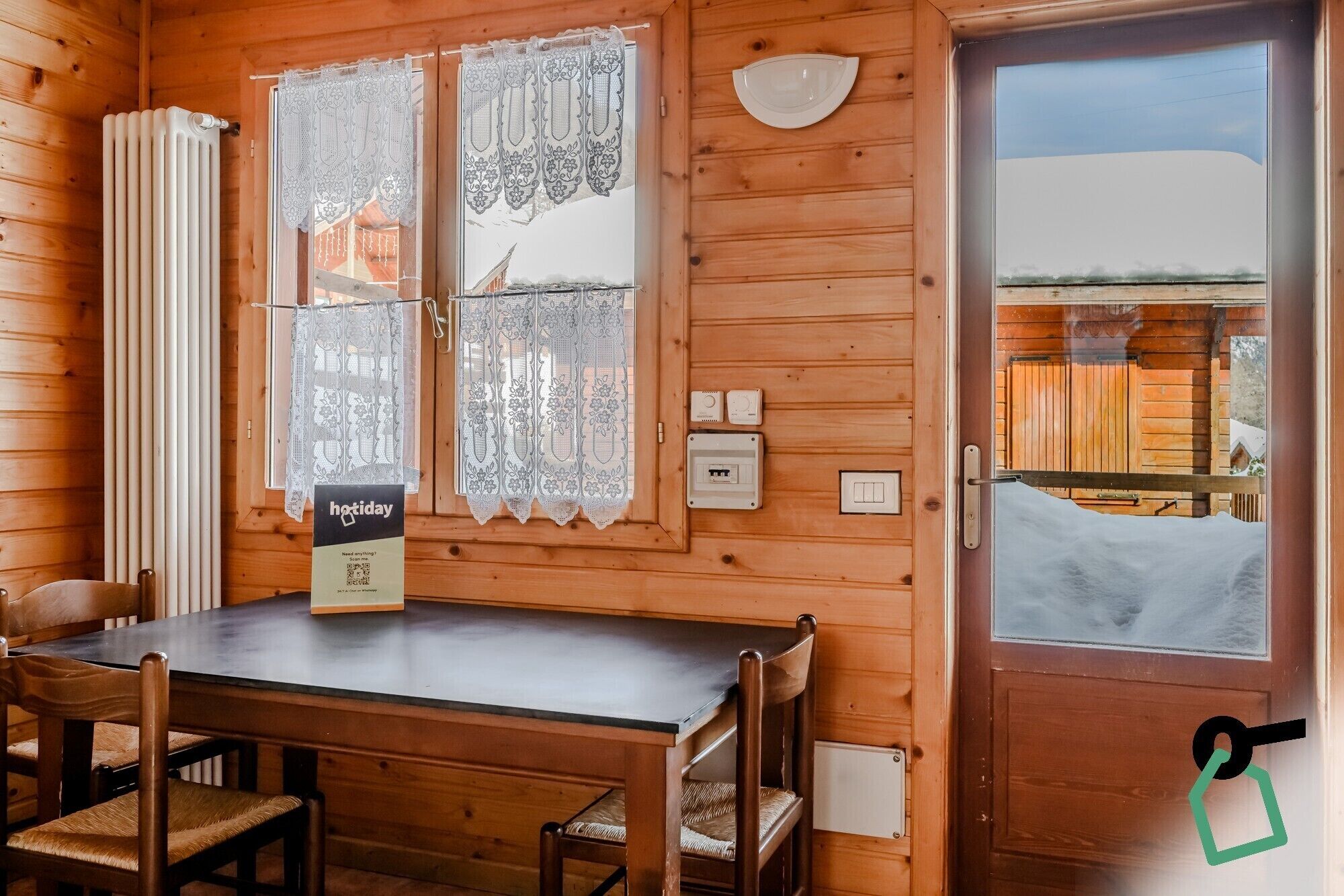 Una cabaña de madera con comedor, cortinas de encaje, un termostato y vista a una estructura de madera en el exterior.