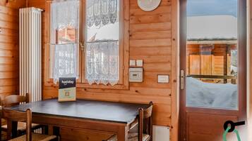 Una cabaña de madera con comedor, cortinas de encaje, un termostato y vista a una estructura de madera en el exterior.