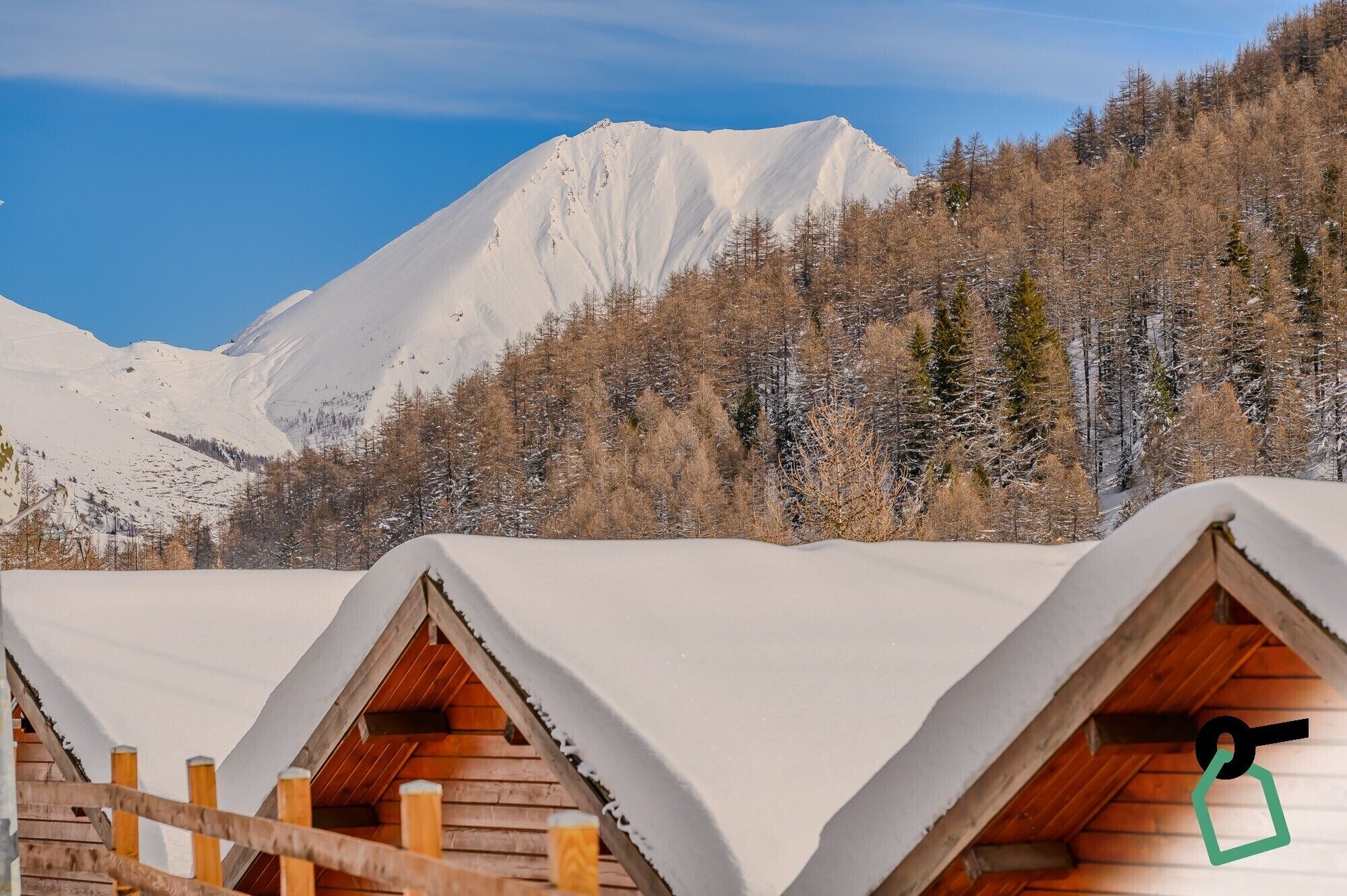 A row of wooden cabins with pitched roofs, set against a backdrop of snow-covered mountains and a clear sky.