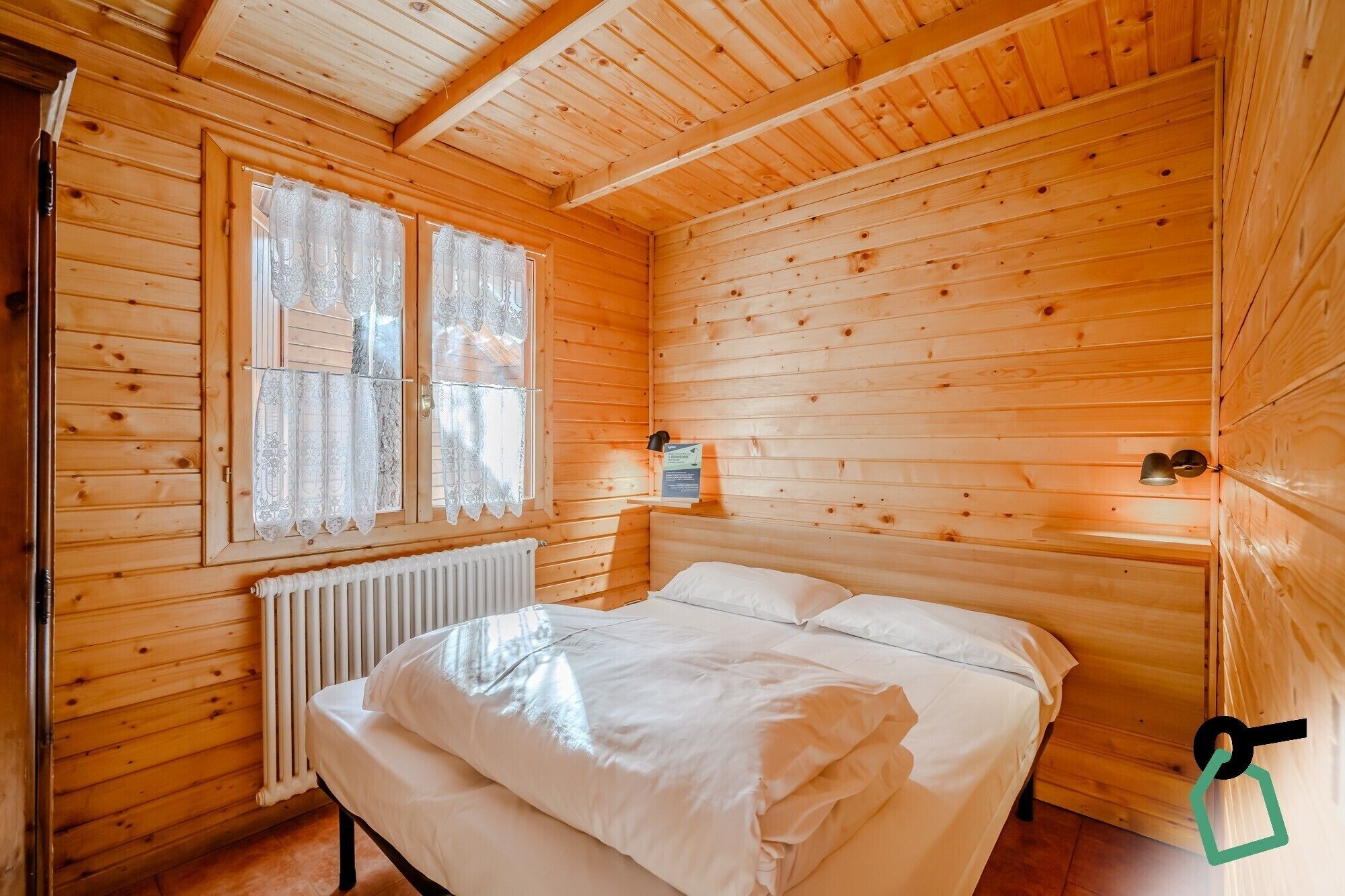 Una habitación de cabaña de madera con una cama, una ventana con cortinas de encaje, un radiador y una lámpara fijada a la pared.