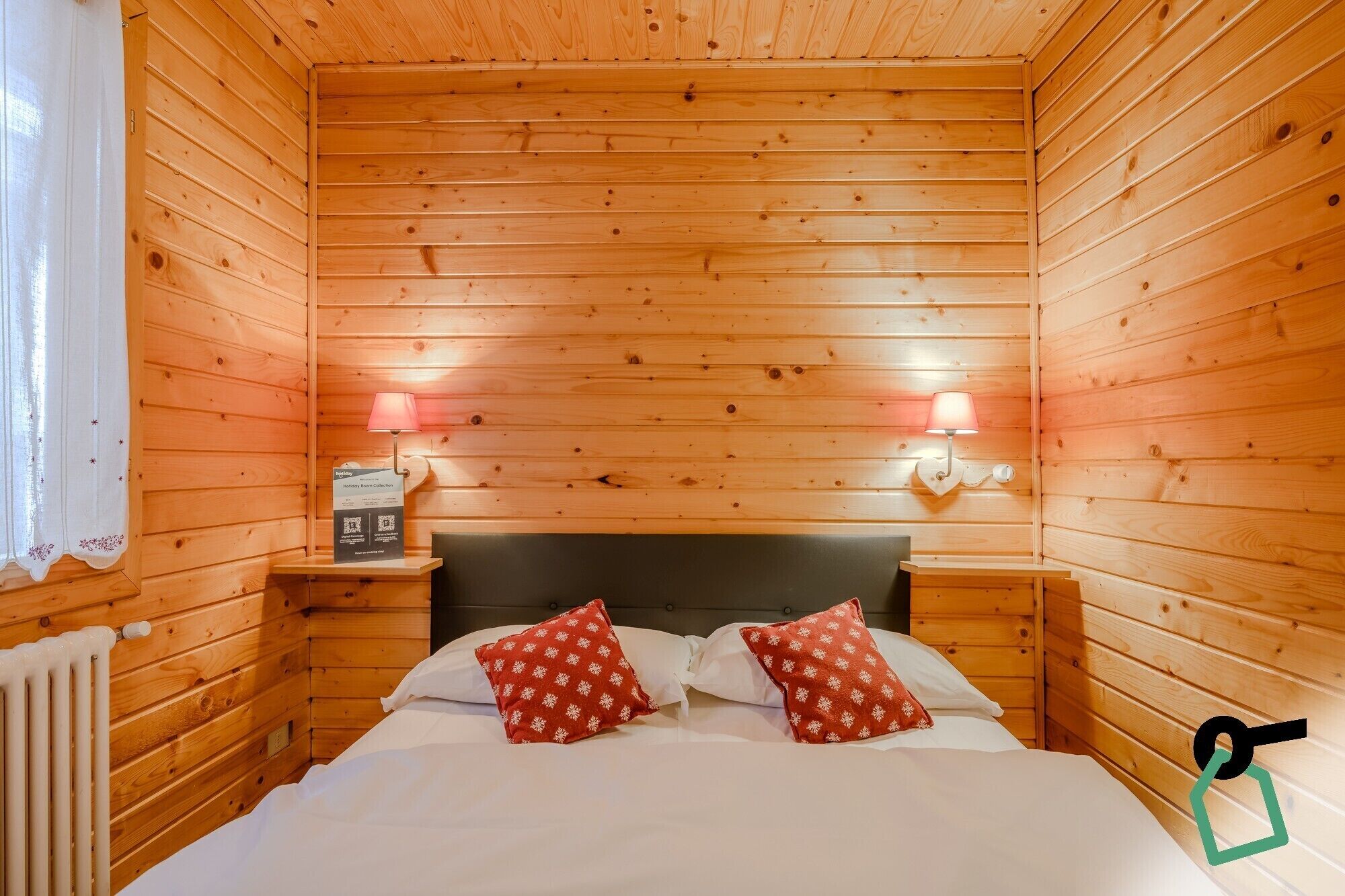 Una habitación acogedora tipo cabaña de madera con una cama, dos mesitas de noche y un letrero que indica una colección de habitaciones para festivos.