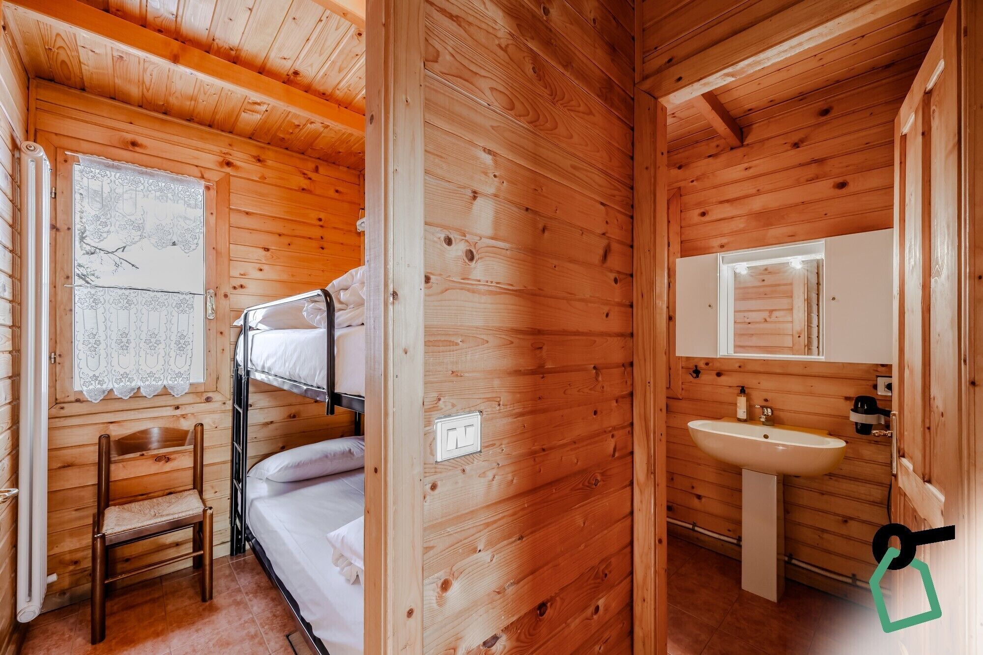 Una habitación de cabaña de madera con literas, una silla, un ventanal con cortinas de encaje y un baño con lavabo y espejo.