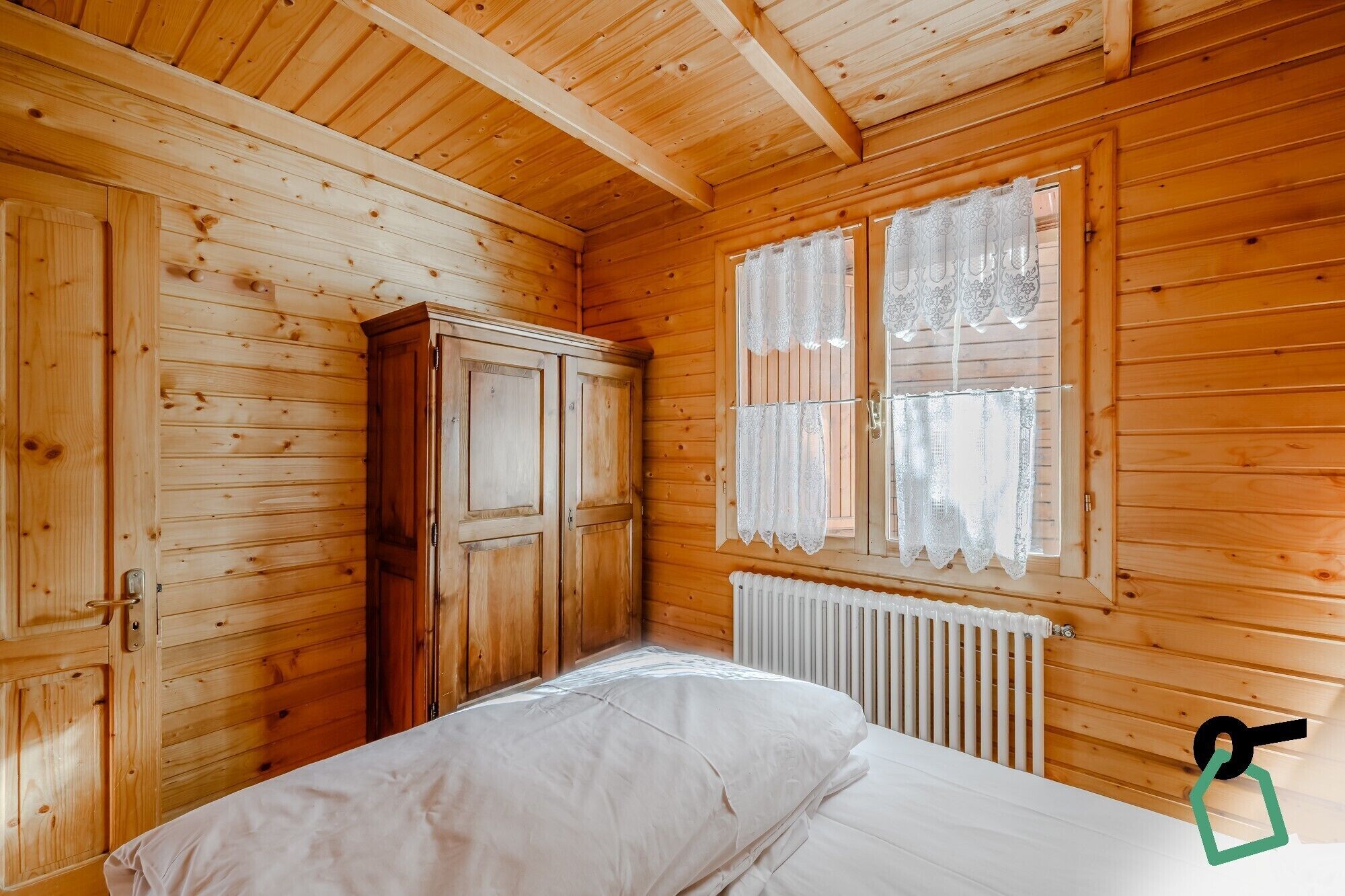 Une chambre dans un chalet en bois, avec un lit, une armoire et une fenêtre ornée de rideaux en dentelle.
