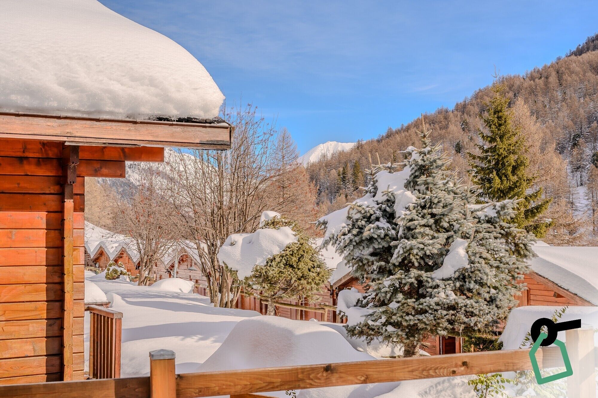 A wooden cabin with a snow-covered roof, surrounded by snow-laden trees and a mountainous backdrop.