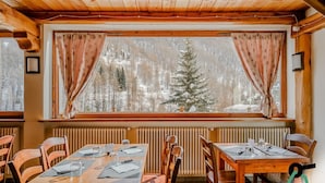 A wooden interior with a dining table set for four, flanked by wooden chairs. A window with curtains offers a view of a snowy forest. - Hotiday Pragelato (Pragelato)