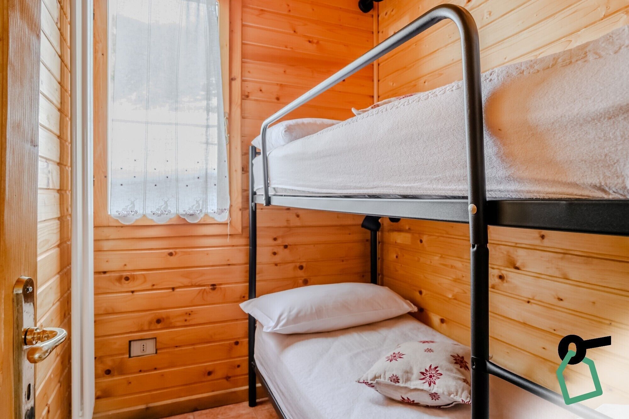 Uma cama beliche com roupa de cama branca, uma parede de madeira e uma janela com cortina de renda.