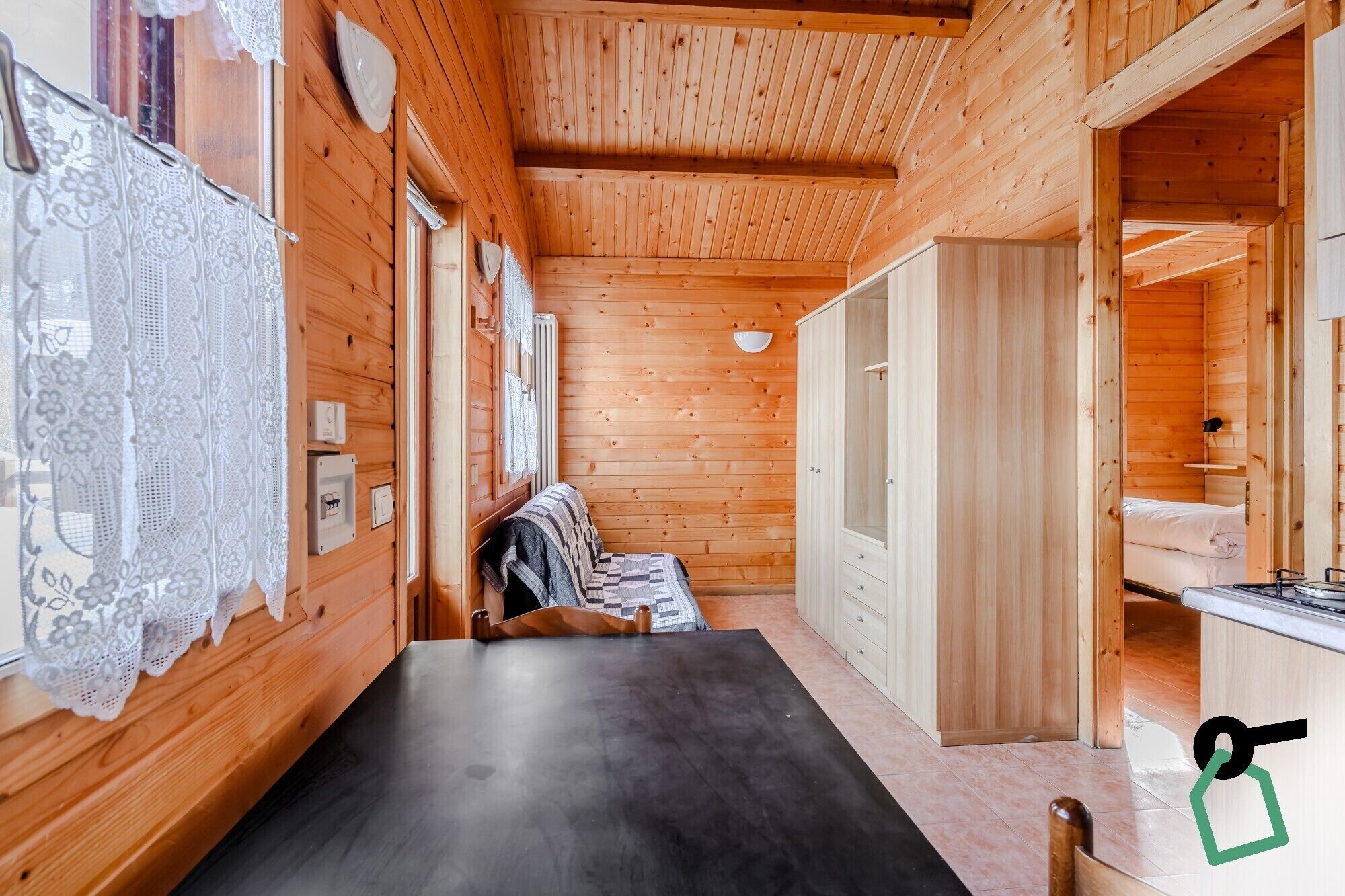 Interior de una cabaña de madera con una mesa de comedor, una cama y un pequeño rincón de cocina.