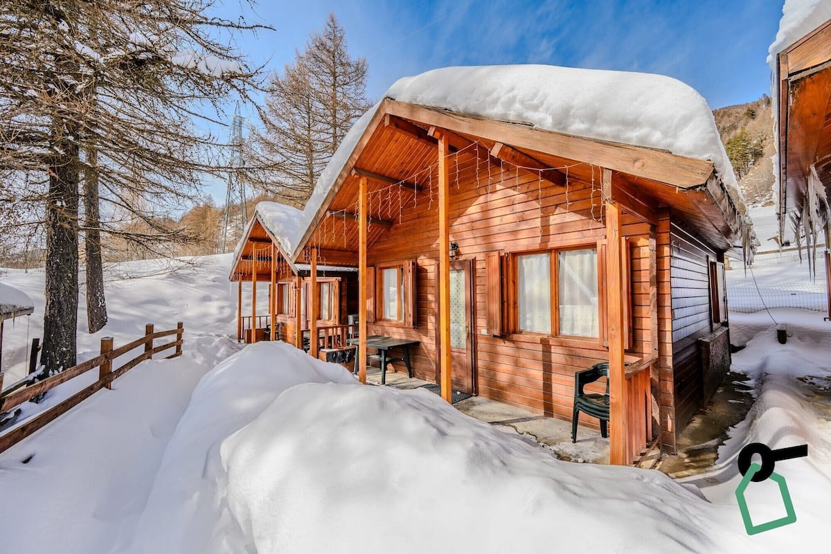 A wooden cabin with a covered porch and a snow-covered roof, surrounded by snow and trees.