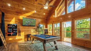 Ein Holzinterieur mit Billardtisch, einem an der Wand befestigten Fernseher und einem großen Fenster mit Blick ins Freie.