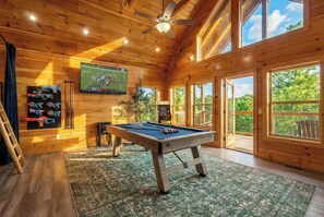 A wooden interior with a pool table, a mounted TV, and a large window offering a view of the outdoors. - Indoor Pool | Great Location | Hot Tub | Arcades (Sevierville)