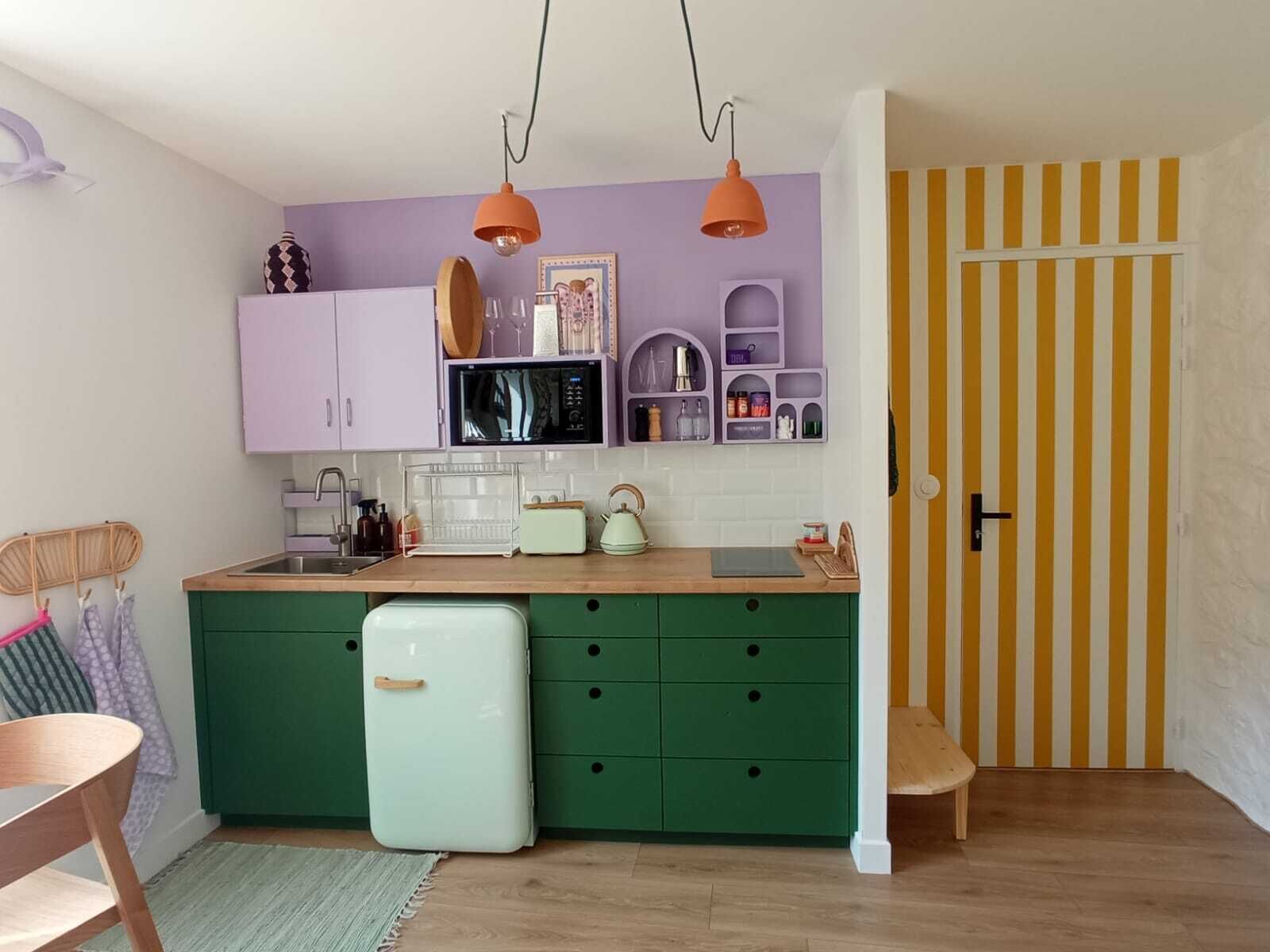 Une cuisine de taille réduite avec des meubles verts, un réfrigérateur blanc et un four à micro-ondes. On y trouve deux lampes suspendues, une chaise en bois et une porte à rayures.