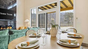 Un comedor con una mesa de madera dispuesta para cuatro, ventanas amplias, una chimenea y un sofá tapizado en tono verde.