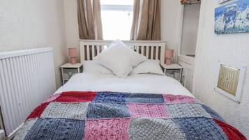 Un dormitorio con una cama blanca, una colcha multicolor de retazos, dos lámparas de noche, una ventana con cortinas y un radiador.