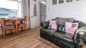 Ein gemütliches Wohnzimmer mit einem Ledersofa, einem Holzesstisch und Stühlen. An der Wand befinden sich Bilderrahmen und durch ein Fenster ist ein Blick ins Freie möglich.