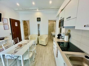 A compact kitchen with white cabinets, a built-in oven, and a sink. There is a dining area with a table and chairs, and a small seating area with two chairs. - Protea house 1 - Alghero (Alghero)