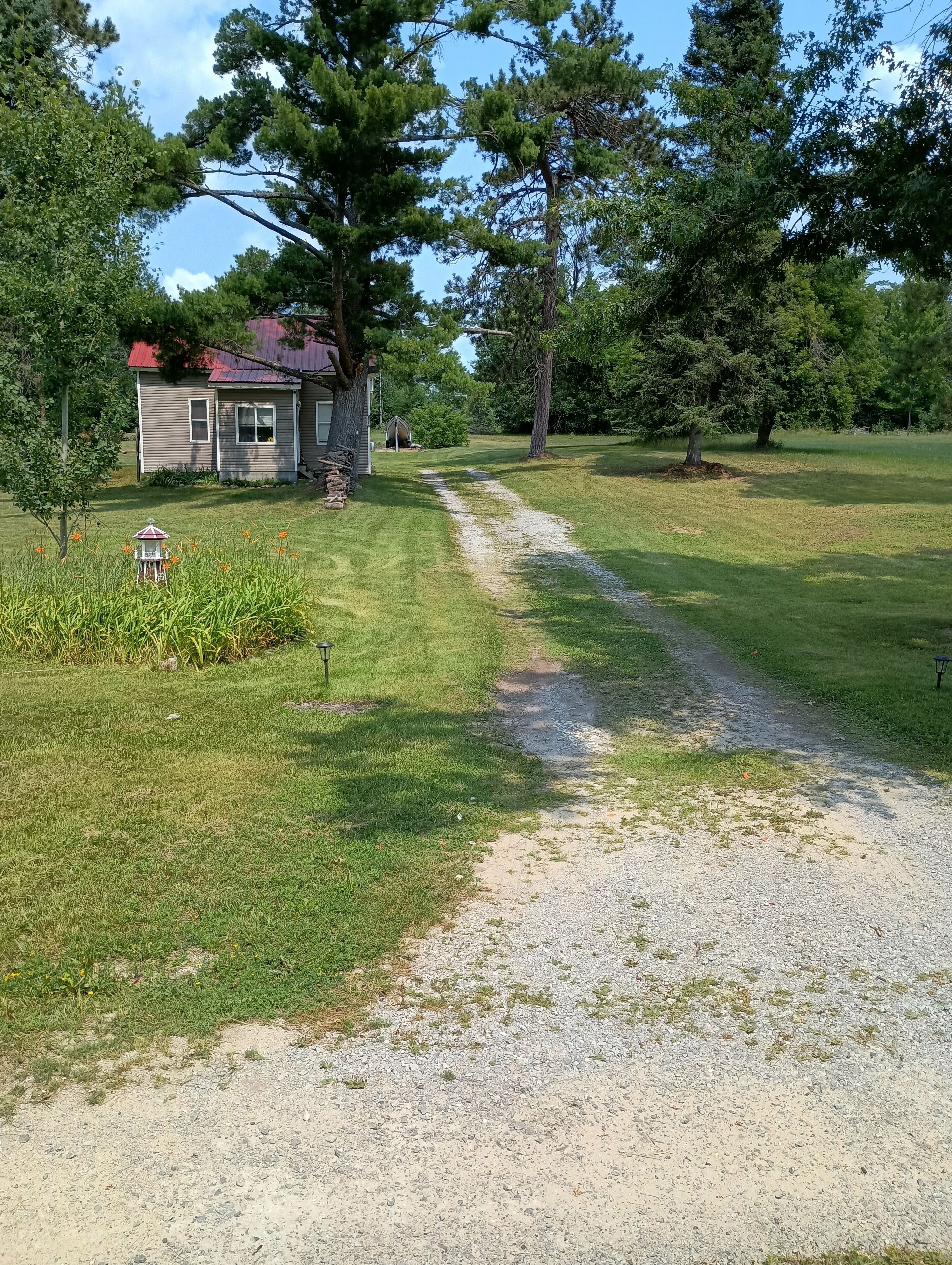 Un chemin de gravier mène à une maison avec un toit rouge, entourée d’arbres et d’une pelouse.