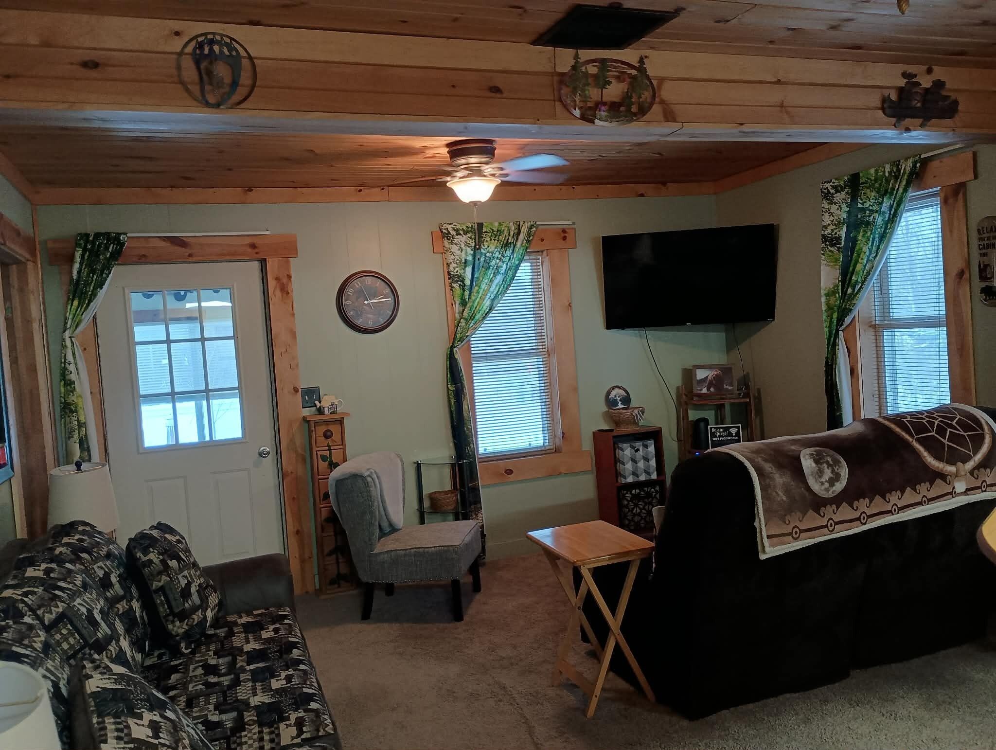 L’intérieur d’un chalet accueillant, avec un plafond en bois, un canapé, un fauteuil, une petite table, un téléviseur et une fenêtre avec des rideaux.