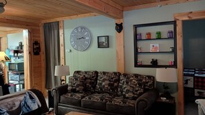 Un salon chaleureux avec un canapé à motifs, un plafond en bois, une grande horloge murale et une étagère ornée de bocaux et d’objets décoratifs.