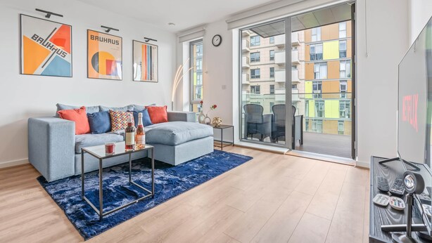 A modern living room with a grey sofa, blue rug, and a large window offering a view of a multi-story building. - Wembley Stadium | Luxury London Apartment | Balcony Views, Netflix, Pool Table! (Wembley)