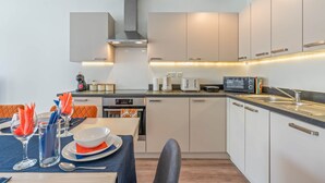 A modern kitchen with white cabinets, a black countertop, and a built-in oven. There is a dining area with a table set for two, and a microwave on the countertop. - Wembley Stadium | Luxury London Apartment | Balcony Views, Netflix, Pool Table! (Wembley)