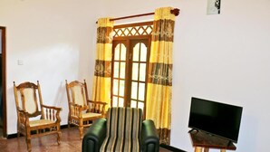 A living room with a sofa, two wooden chairs, a television on a wooden stand, and a window with curtains. - lavish eco jungle (Weerawila)