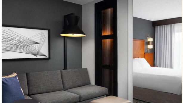 Quarto de hotel moderno com sofá cinzento, uma mesinha redonda, um banquinho de madeira, um abajur de chão preto e uma cama com roupa de cama branca.